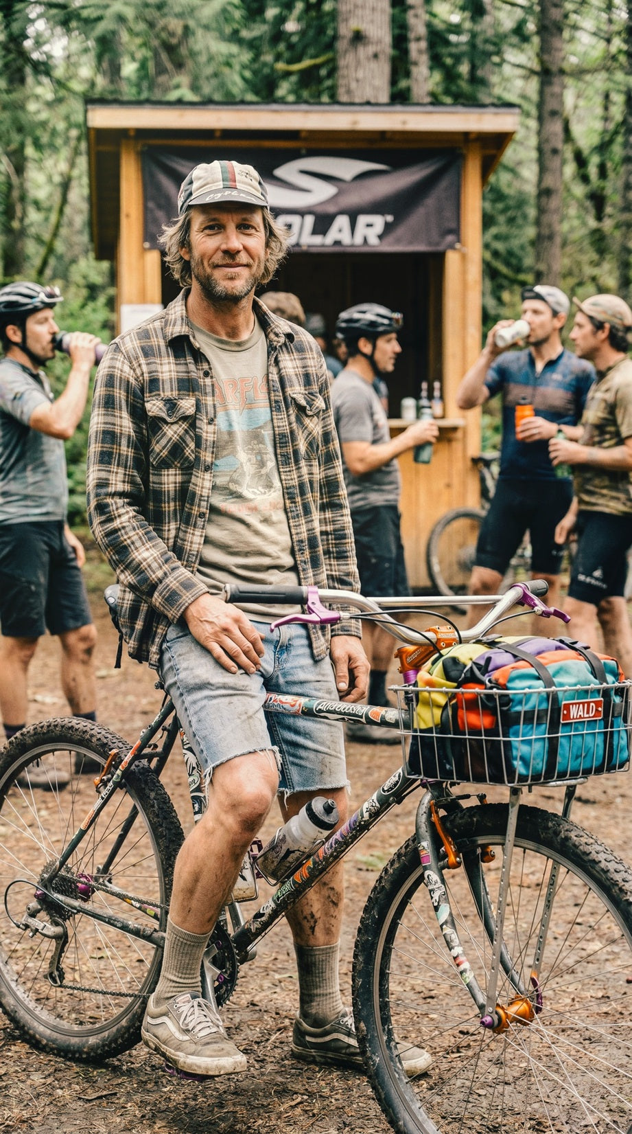Alternative dirtbag cyclist posing with a custom steel mountain bike at a grassroots cycling gathering. Features wide alt-bars, Wald basket, colorful bikepacking gear, vintage flannel shirt, denim jorts, and DIY bike modifications. Outdoor cycling community, tracklocross culture, bikepacking lifestyle, steel bike adventure, party pace riding, forest cycling event.