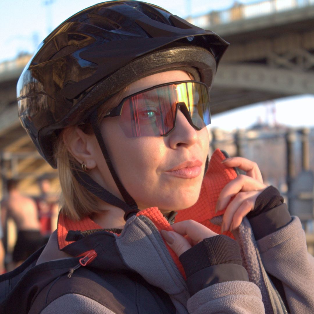 Woman wearing SAOLAR Emberline photochromic sports sunglasses with sleek red-orange reflective lenses and dark frame, under evening light near a bridge.