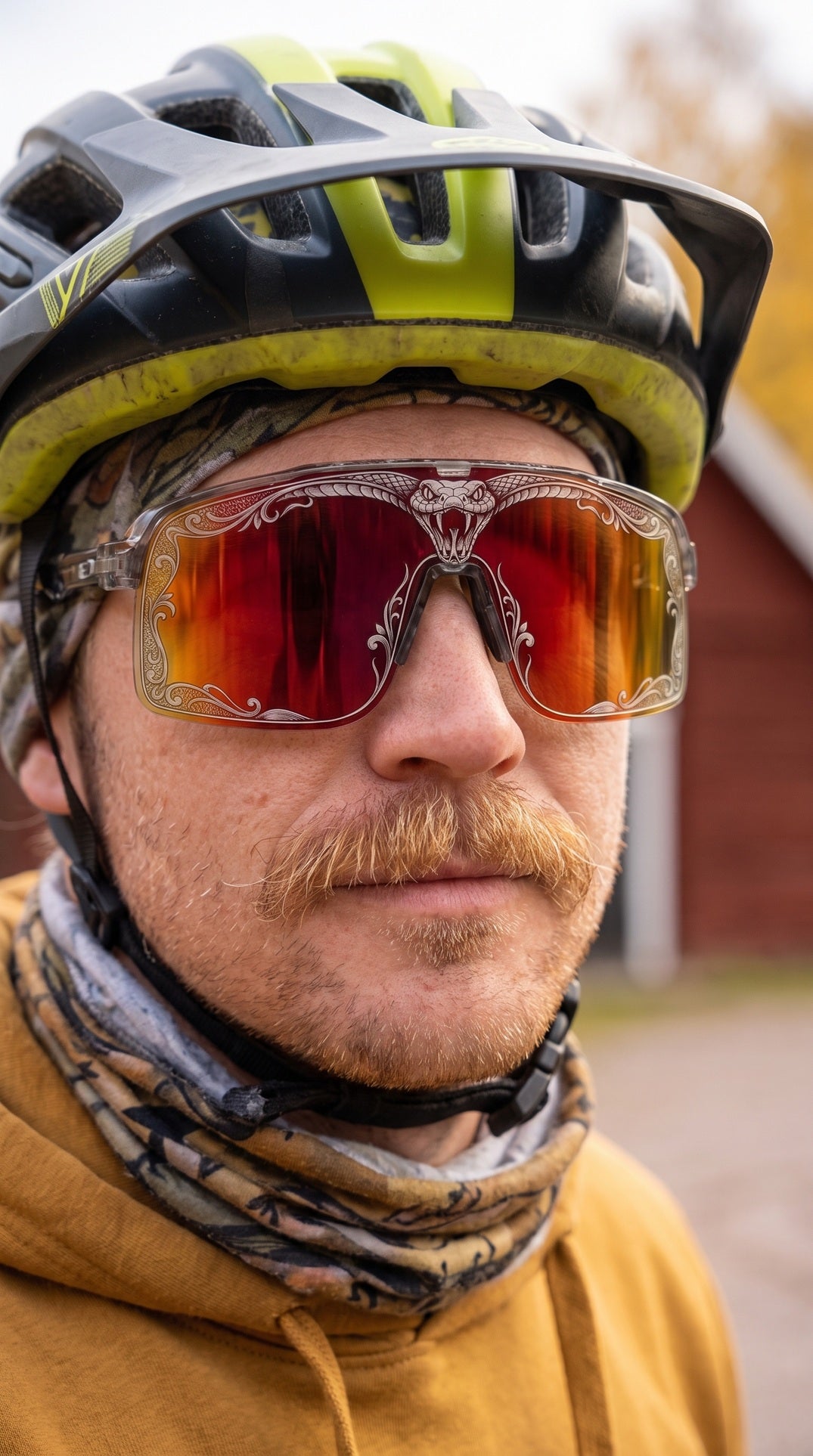 Close-up portrait of moustached cyclist wearing SAOLAR Flareforce Venom sunglasses with red mirrored lenses, snake artwork, grey helmet and mustard hoodie.