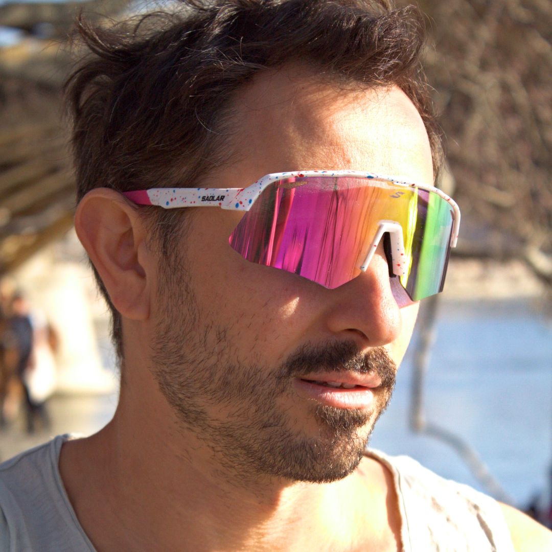 Man wearing SAOLAR Flicker photochromic sports sunglasses with vibrant pink-to-green mirrored lens and white speckled frame, in sunlight outdoors.