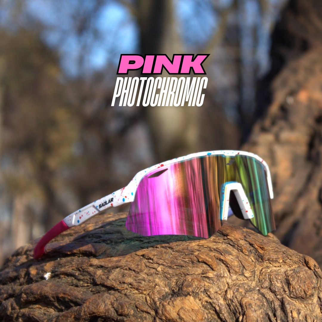 SAOLAR Flicker pink photochromic sunglasses with a white splattered frame, wraparound lens, and pink temples, displayed on rugged wood outdoors.