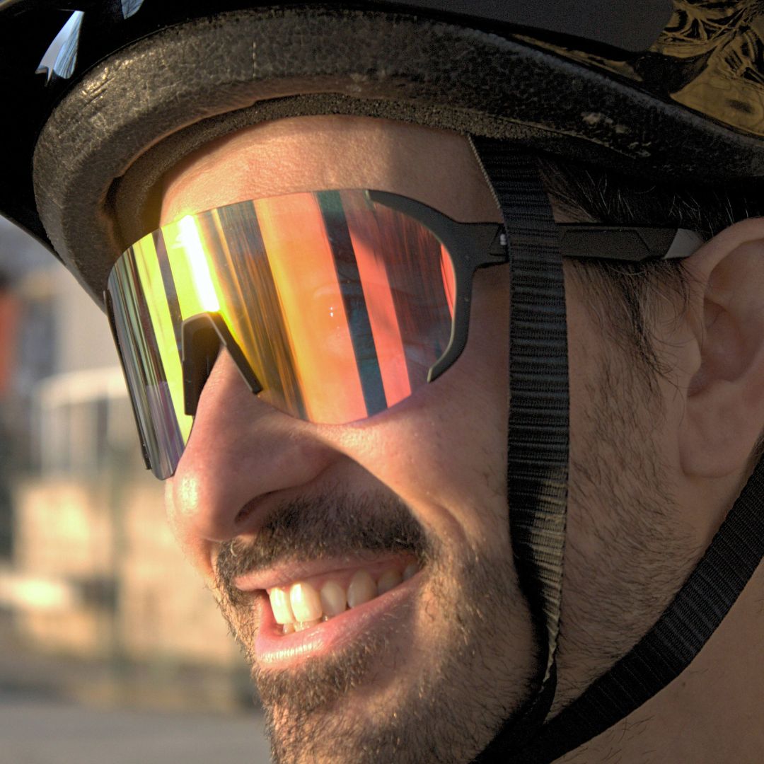 Close-up of a smiling man wearing SAOLAR Hyperion photochromic sports sunglasses with orange-gold mirrored lenses and matte black frame, under bright sunlight.