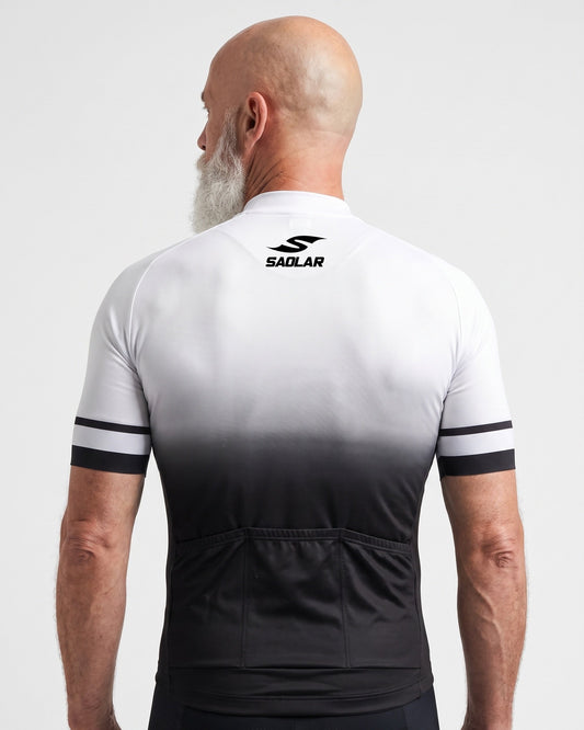 Back studio view of a mature male cyclist wearing the SAOLAR Gradient cycling jersey. The white-to-black fade extends from the shoulders to the waist, with striped sleeve bands, the SAOLAR logo below the collar, and three practical rear storage pockets clearly visible above black cycling bib shorts on white backdrop.
