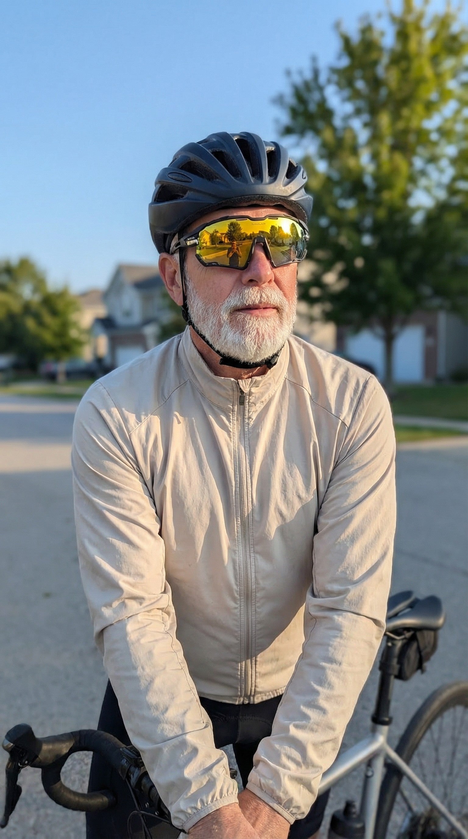 Senior cyclist wearing SAOLAR mirrored sunglasses riding calmly on suburban road, morning light, helmet and beige jacket, comfortable everyday cycling eyewear lifestyle scene.