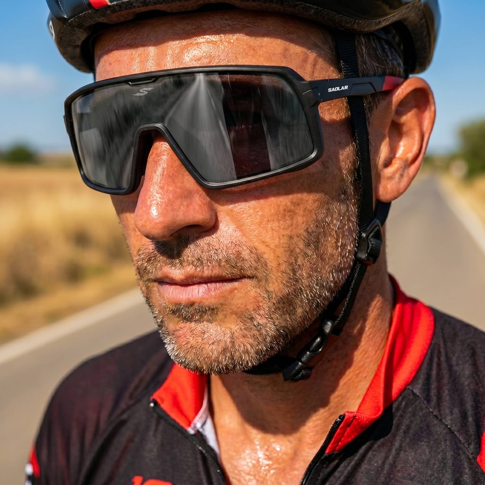 Close-up portrait of a male cyclist wearing SAOLAR Shadelane Classic sunglasses with dark photochromic lenses, riding on a sunlit road. The lightweight wraparound frame provides full eye coverage, reducing glare and enhancing contrast. Sweat and helmet details emphasize performance, comfort, and durability for long-distance cycling in bright outdoor conditions daily.