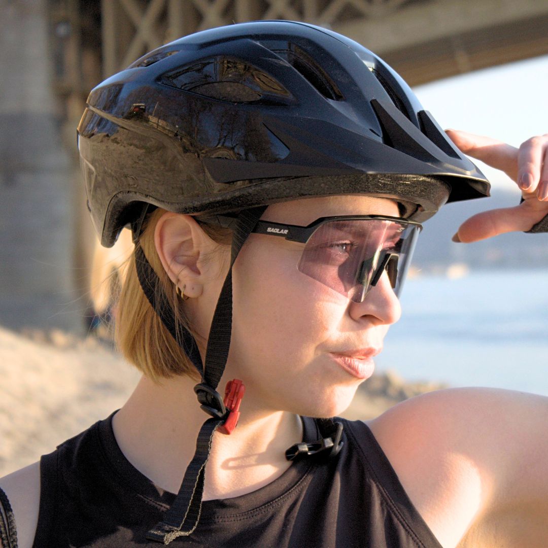 Woman wearing SAOLAR Stratos photochromic sports sunglasses and bike helmet, looking into sunlight near a bridge and waterfront.