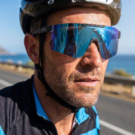 Close-up of male cyclist wearing SAOLAR Tundrake photochromic full revo blue sports sunglasses, reflective shield lens adapting to sunlight, lightweight ergonomic frame, UV400 protection, panoramic vision, ideal for road cycling performance, reducing glare, enhancing clarity, modern athletic design, breathable comfort, outdoor endurance riding along coastal road under bright daylight conditions