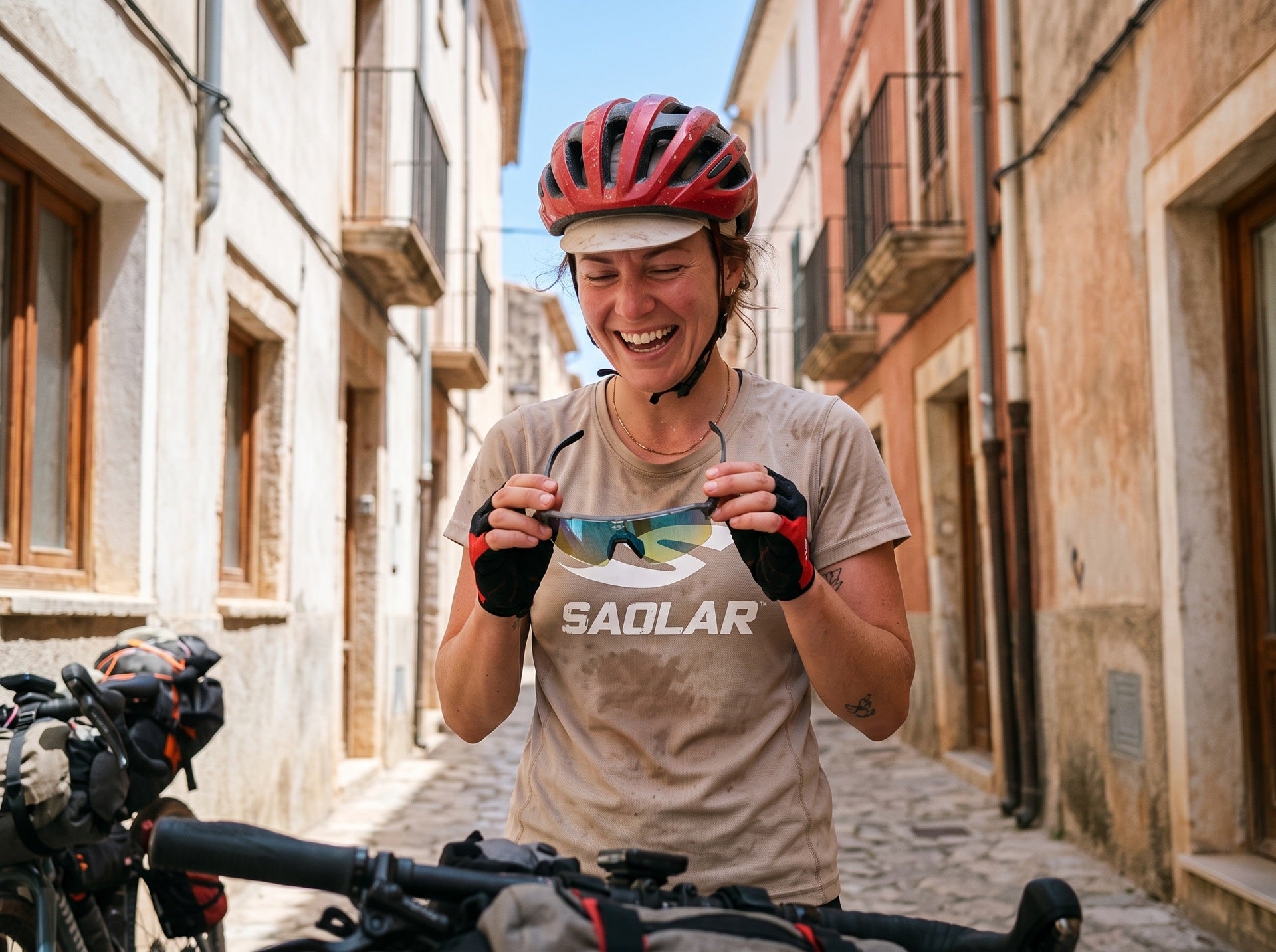 Smiling female cyclist holding SAOLAR Flareforce sunglasses during a bikepacking stop on a charming European cobblestone street. Wearing a red cycling helmet and dusty SAOLAR jersey, she laughs while admiring the mirrored photochromic lenses. Loaded touring bikes frame the foreground, capturing an authentic outdoor cycling adventure in warm summer sunshine.